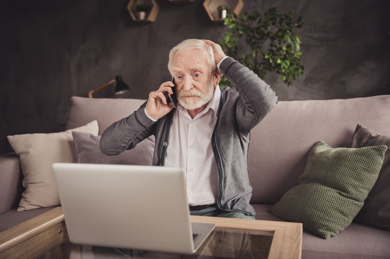 Senior sitzt mit Handy am Ohr in einem Wohnzimmer auf dem Sofa und fasst sich mit der freien Hand an den Kopf. Er ist offensichtlich erschrocken.